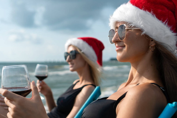 Dwie młode szczęśliwe kobiety w strojach kąpielowych i kapelusze Świętego Mikołaja z kieliszkiem wina w rękach siedzą na składanych krzesłach nad morzem i cieszą się chwilą. Święta Bożego Narodzenia w ciepłych krajach. - Zdjęcie, obraz