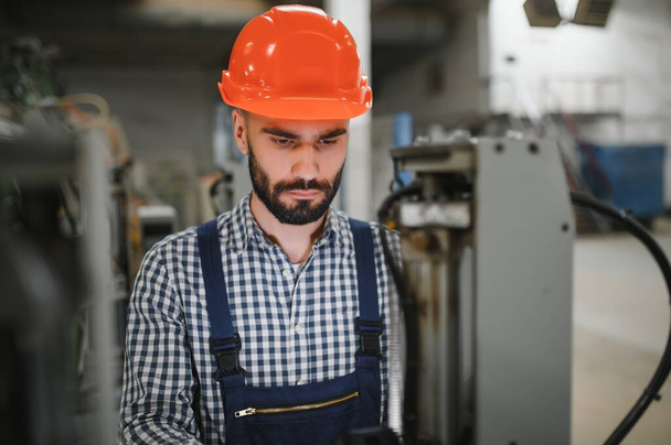 Portrét profesionálního inženýra těžkého průmyslu, pracovníka v bezpečnostní uniformě, tvrdý klobouk s úsměvem. V pozadí Rozostřená velká průmyslová továrna - Fotografie, Obrázek