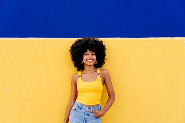 Beautiful young happy african woman with afro curly hairstyle strolling in the city - Cheerful black student portrait on colorful wall background - Фото, изображение