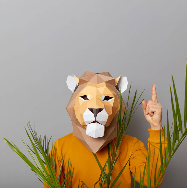 Indoor shot of man wearing lion paper mask standing isolated over gray background, pointing finger up, showing copy space for advertisement or promotional text. - Fotografie, Obrázek
