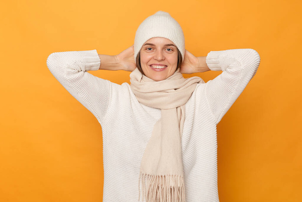 Indoor shot of cheerful joyful positive woman wears white sweater, scarf and cap, standing with hands on behind her head, looking at camera with toothy smile, standing isolated over yellow background. - Foto, afbeelding