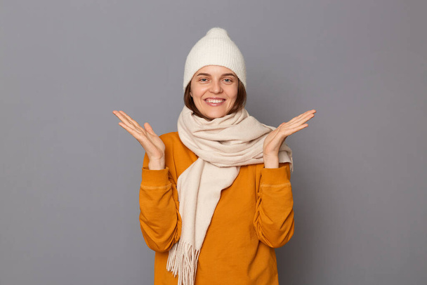 Image of extremely happy overjoyed woman wearing orange sweater, scarf and cap standing isolated over gray background, raised her arms, being in very good mood. - Φωτογραφία, εικόνα