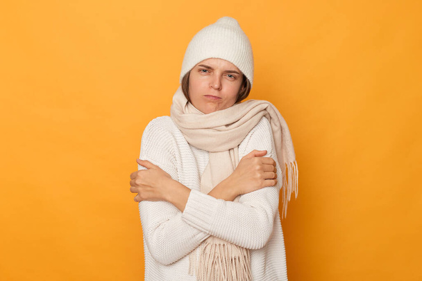 Portrait of sick freeze woman wearing white sweater, scarf and cap standing isolated over yellow background, hugging herself, trying to get warm, expressing sadness, feels cold. - Fotoğraf, Görsel