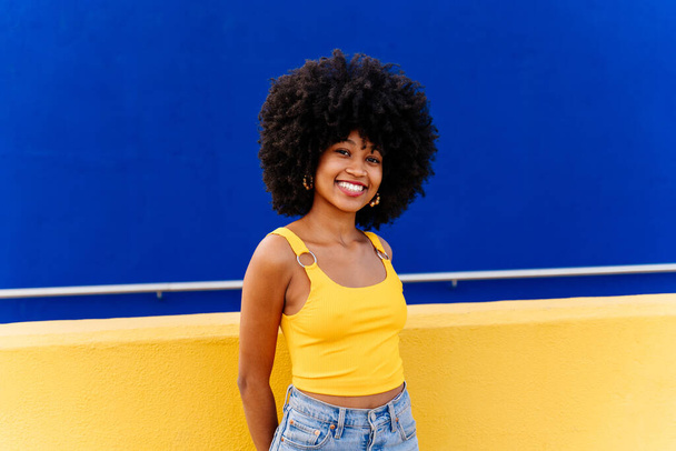 Beautiful young happy african woman with afro curly hairstyle strolling in the city - Cheerful black student portrait on colorful wall background - 写真・画像