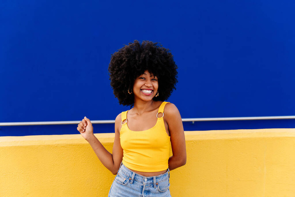 Beautiful young happy african woman with afro curly hairstyle strolling in the city - Cheerful black student portrait on colorful wall background - Foto, Imagem