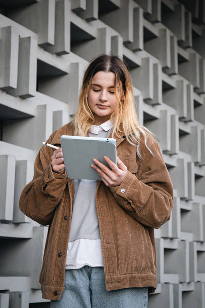 Jolie jeune femme dans une veste en velours côtelé dessine avec un crayon sur une tablette numérique, sur un fond flou d'un mur géométrique en béton dans la ville.. - Photo, image