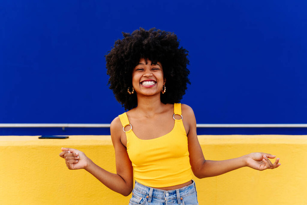 Beautiful young happy african woman with afro curly hairstyle strolling in the city - Cheerful black student portrait on colorful wall background - Zdjęcie, obraz