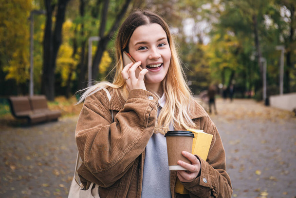 Atrakcyjna młoda studentka w sztruksowej kurtce, z książką i filiżanką kawy rozmawia przez telefon w jesiennym parku.. - Zdjęcie, obraz