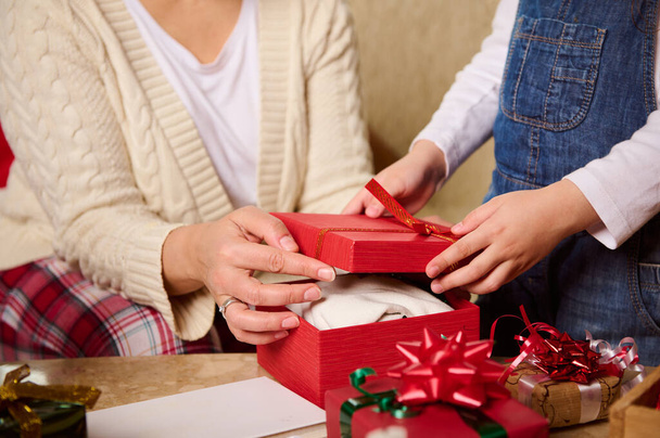 Szczegóły: dłonie kobiety - mamy i dziecka - córki pakujące prezenty w piękne, stylowe, czerwone pudełko. Szczęśliwej magicznej atmosfery Bożego Narodzenia w domu. Przygotowania do Nowego Roku. Zimowe wakacje atmosfera - Zdjęcie, obraz