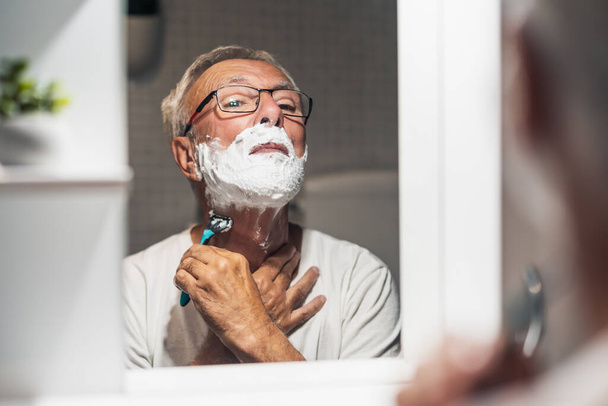 Senior man is shaving his beard with razor and shaving foam in bathroom. - Foto, afbeelding