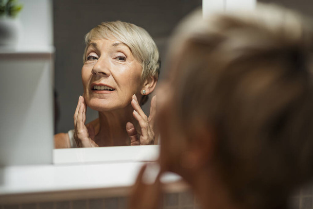 Зрелая женщина смотрит на свое лицо в ванной. Она исследует морщины на коже.. - Фото, изображение