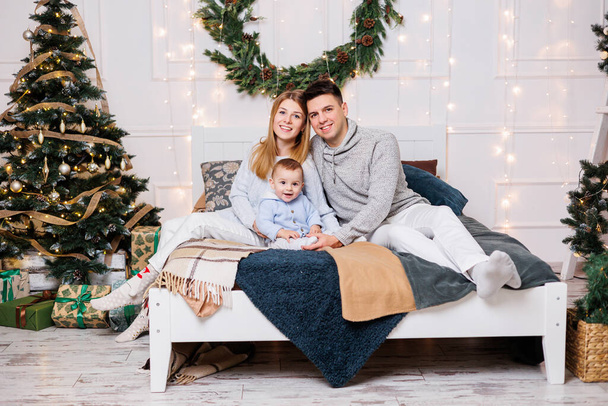 A cheerful young couple with a little son are playing on the bed near the Christmas tree. New Year's interior in the bedroom. Christmas tree with toys. Festive family atmosphere. - Фото, зображення