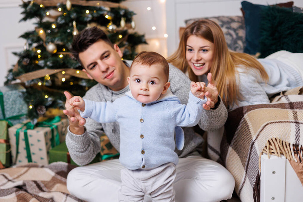 Leuke jongen met gelukkige ouders bij de kerstboom. Een familie met een klein kind in een slaapkamer in de buurt van een versierde kerstboom. Feestelijke nieuwjaarssfeer. Familie viering - Foto, afbeelding