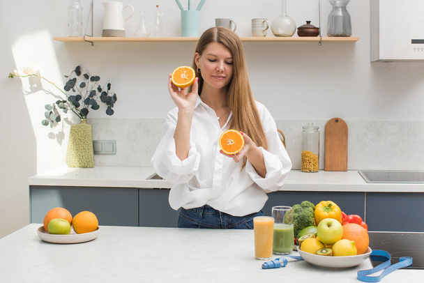 A young blond woman holds orange halves in her hands in the kitchen next to a table with fruits. The concept of diet, healthy eating. - Fotografie, Obrázek