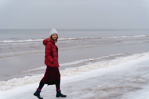 Een vrouw in rode warme kleren wandelt in de winter langs de zee. - Foto, afbeelding