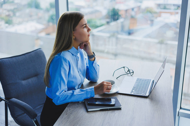 Piękna bizneswoman w niebieskiej koszuli, siedząca w biurze z laptopem i robiąca interesy. Nowoczesne biuro w wieżowcu z oknami. - Zdjęcie, obraz