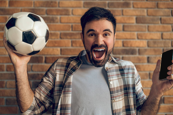 Portrait of an emotional guy holding the football and cellphone in the lifted hands and yelling - Foto, afbeelding