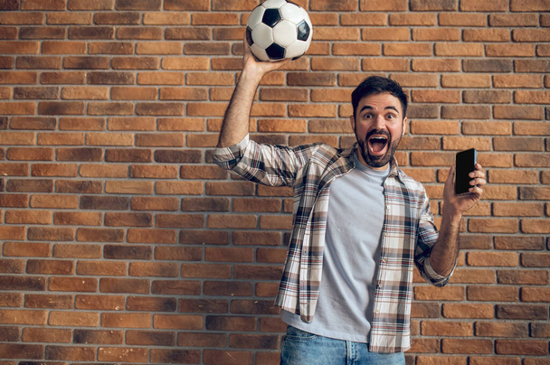 Emotional soccer buff posing for the camera against the brick wall with the ball and smartphone - Fotoğraf, Görsel