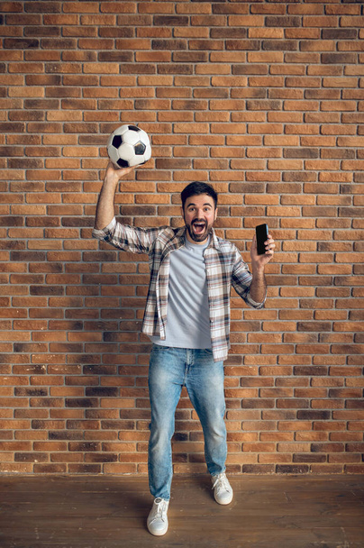 Full-size portrait of a screaming guy holding the soccer ball and mobile phone in the lifted hands - Φωτογραφία, εικόνα