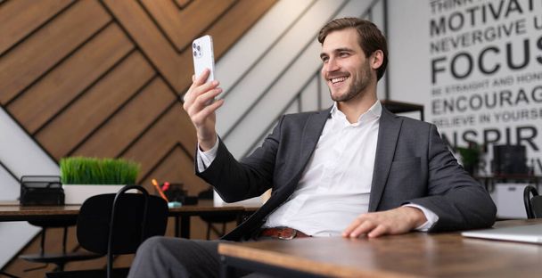 молодой бизнесмен, сидящий за столом в офисе и разговаривающий по видеосвязи с помощью мобильного телефона. - Фото, изображение