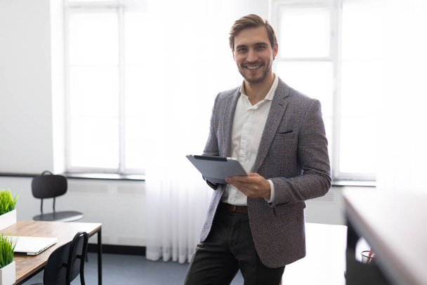 彼の手に電子タブレットを持つ若いビジネスマンは,オフィスの真ん中に笑顔で立っています. - 写真・画像