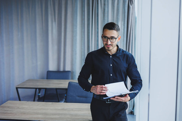 A Caucasian business man in glasses and a black shirt sits in the office and looks at sales growth charts. A young successful businessman in classic clothes stands by the window in the office - Foto, Imagen
