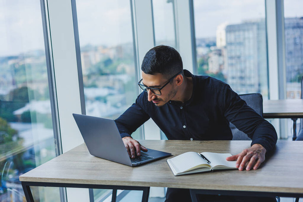 Homme d'affaires prospère en lunettes et chemise noire assis à un bureau et travaille sur un projet via un ordinateur portable dans un bureau moderne, en regardant l'écran. - Photo, image