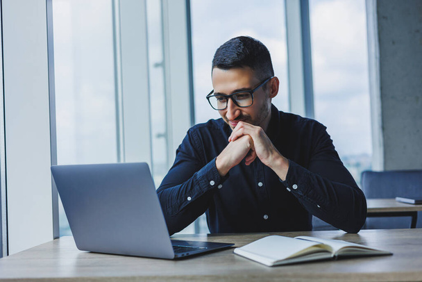 Sikeres férfi üzletember szemüvegben és fekete ingben ül az asztalnál, és egy projekten dolgozik egy laptopon keresztül egy modern irodában, a képernyőt bámulva.. - Fotó, kép