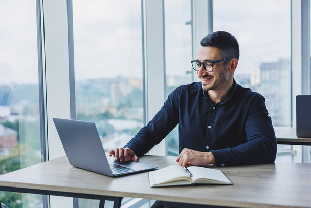 Młody, odnoszący sukcesy pracownik płci męskiej w okularach i koszulce siedzący przy biurku i pracujący nad projektem za pośrednictwem laptopa w nowoczesnym biurze patrzący na ekran - Zdjęcie, obraz