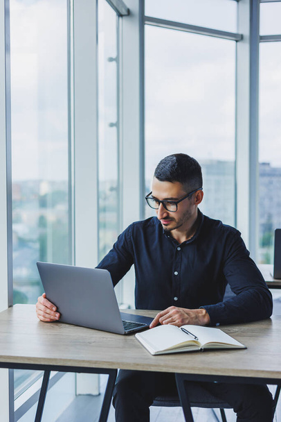 Młody, odnoszący sukcesy pracownik płci męskiej w okularach i koszulce siedzący przy biurku i pracujący nad projektem za pośrednictwem laptopa w nowoczesnym biurze patrzący na ekran - Zdjęcie, obraz
