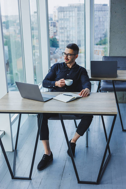 Ein gutaussehender Manager kaukasischen Aussehens sitzt in einem schwarzen Hemd an einem Tisch und trinkt Kaffee in einem modernen Büro. Arbeitsatmosphäre im Büro. - Foto, Bild