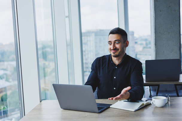 Ένας επιτυχημένος επιχειρηματίας με λευκή εμφάνιση με μαύρο πουκάμισο κάθεται σε ένα γραφείο χρησιμοποιώντας ένα φορητό υπολογιστή σε ένα σύγχρονο γραφείο. Η εργασιακή ατμόσφαιρα στο γραφείο. - Φωτογραφία, εικόνα
