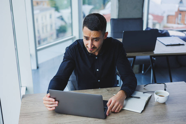 Un uomo d'affari di successo di aspetto caucasico in una camicia nera è seduto a una scrivania utilizzando un computer portatile in un ufficio moderno. Atmosfera di lavoro in ufficio. - Foto, immagini
