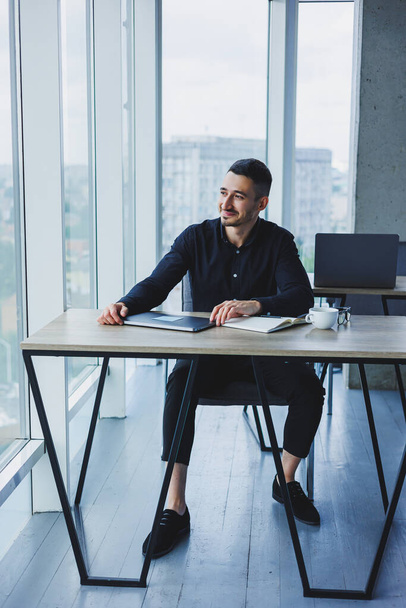 Успешный бизнесмен с кавказской внешностью в черной рубашке сидит за столом, используя ноутбук в современном офисе. Рабочая атмосфера в офисе. - Фото, изображение