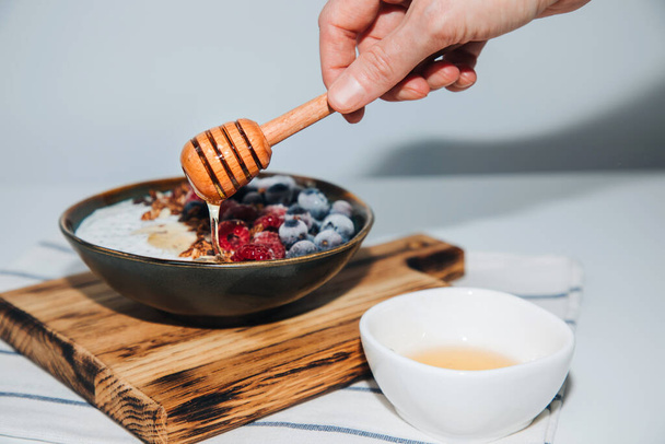 Female hand pours honey yoghurt in green plate with blueberries, raspberries, chia seeds and granola with a wooden board on a kitchen towel. The concept of healthy eating. Front view - 写真・画像