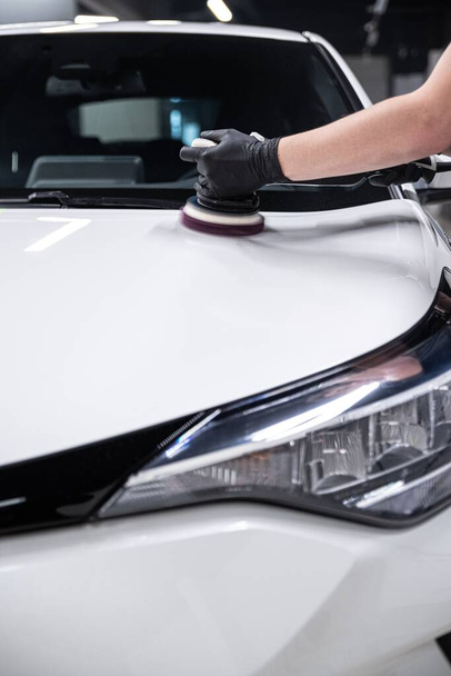 Υπάλληλος πλυντηρίου αυτοκινήτων ή αυτοκινήτου που περιγράφει λεπτομερώς στούντιο γυαλίζει τη βαφή ενός λευκού αυτοκινήτου με ηλεκτρικό γυαλιστή - Φωτογραφία, εικόνα