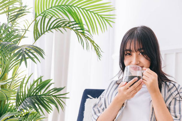 Młoda ładna kobieta uśmiecha się i siedzi na krześle relaksując się pijąc filiżankę gorącej kawy w nowoczesnym salonie z zieloną rośliną, uzdrawiające odbicie w otoczeniu wewnątrz w przytulnym domu. - Zdjęcie, obraz