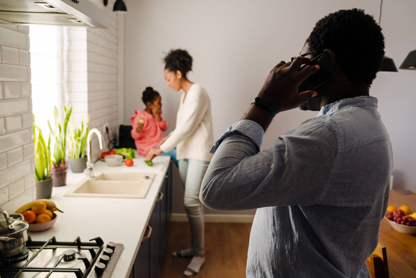 Afrikaans amerikaanse man praten op mobiele telefoon terwijl vrouw en dochtertje het maken van ontbijt in de keuken - Foto, afbeelding