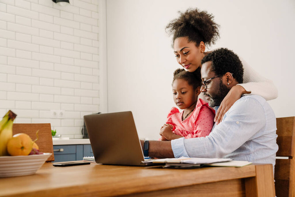 African american family man and woman with little daughter smiling while using laptop in kitchen - Foto, immagini