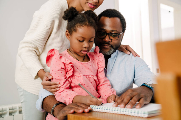 African american little girl writing in notepad with pen while sitting with parents by table at home - Φωτογραφία, εικόνα