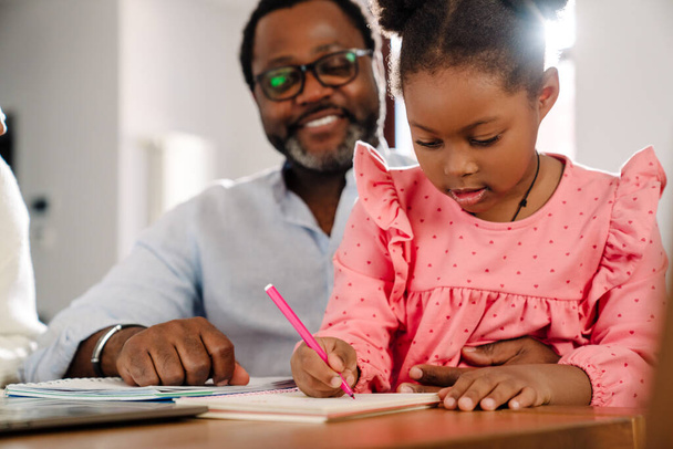 African american little girl drawing in notepad with marker while sitting with parents by table at home - Fotoğraf, Görsel