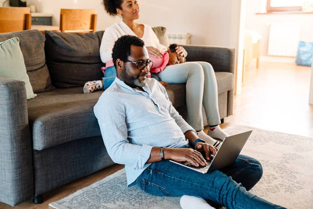 African american woman with little daughter sitting on sofa while man using laptop at home - Foto, afbeelding