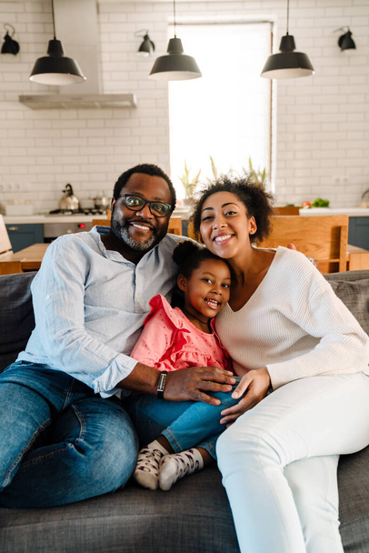 Afrikai amerikai család nő és férfi kislányával mosolyog és otthon ül a kanapén - Fotó, kép
