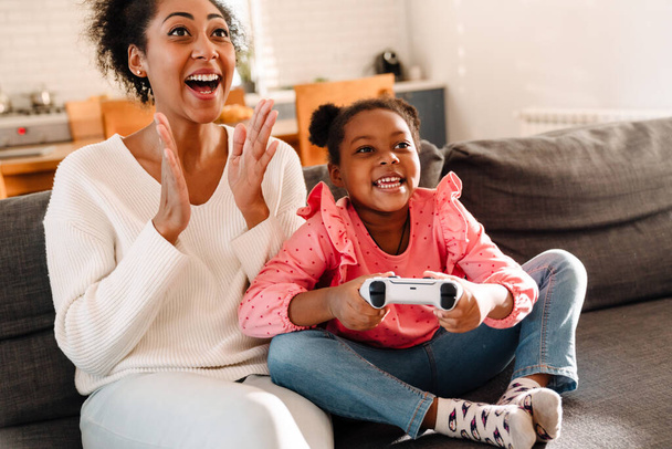 African american woman and little girl sitting on sofa and playing video game with controller at home - Fotografie, Obrázek