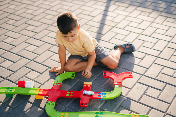 Funny boy 4-5 years old enthusiastically playing with a plastic railroad set, outdoors. Educational toys for preschool and kindergarten children. Training - Photo, Image