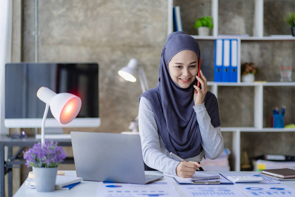 Gelukkig moslim zakenvrouw in hijab op het werk Lachende Arabische vrouw werkt op laptop en praten op smartphone met geld grafiek op de tafel, financiële boekhouding concept - Foto, afbeelding