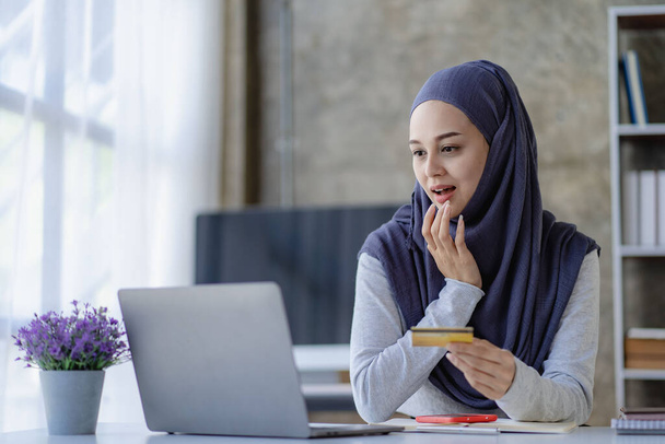 gelukkige jonge moslim vrouw dragen hijab Hand holding debetkaart en online bestellen op laptop zittend op kantoor winkelen en het uitvoeren van transacties op het internet op een bankrekening - Foto, afbeelding