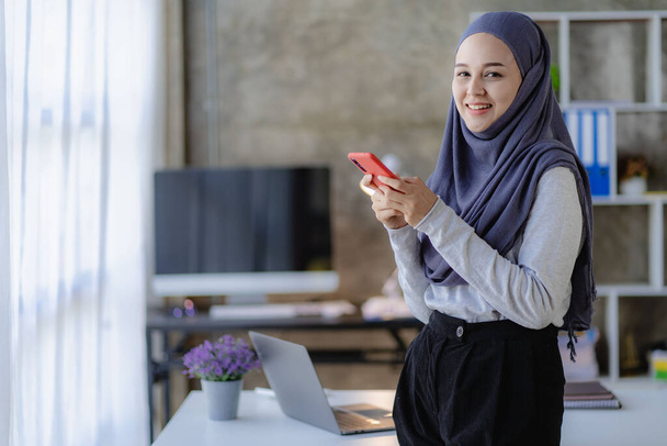 Χαρούμενη Μουσουλμάνα επιχειρηματίας με μαντίλα στη δουλειά Χαμογελώντας Αραβίδα γυναίκα που εργάζεται σε φορητό υπολογιστή και μιλάει σε smartphone με γράφημα χρημάτων στο τραπέζι, έννοια της χρηματοοικονομικής λογιστικής - Φωτογραφία, εικόνα
