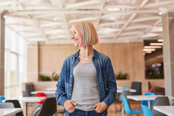 Happy young female in casual clothes with dyed blond hair holding hands in pockets and looking away with smile while, standing on blurred background of workspace - Foto, Bild