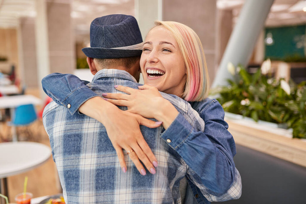 Cheerful blond woman smiling and embracing black boyfriend in checkered shirt and hat during friends meeting in restaurant - Zdjęcie, obraz